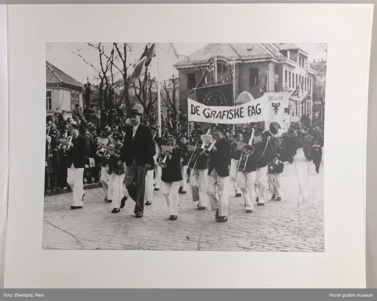 1. mai-tog i Stavanger. En mann og et korps går foran banneret til "De grafiske fag". Den hvite fanen til høyre i bakgrunnen er fanen for Stavanger Grafiske Fagforening (Typografene i Stavanger). Bak banneret "De grafiske fag" skimtes fanen til Bokbindernes fagforening.