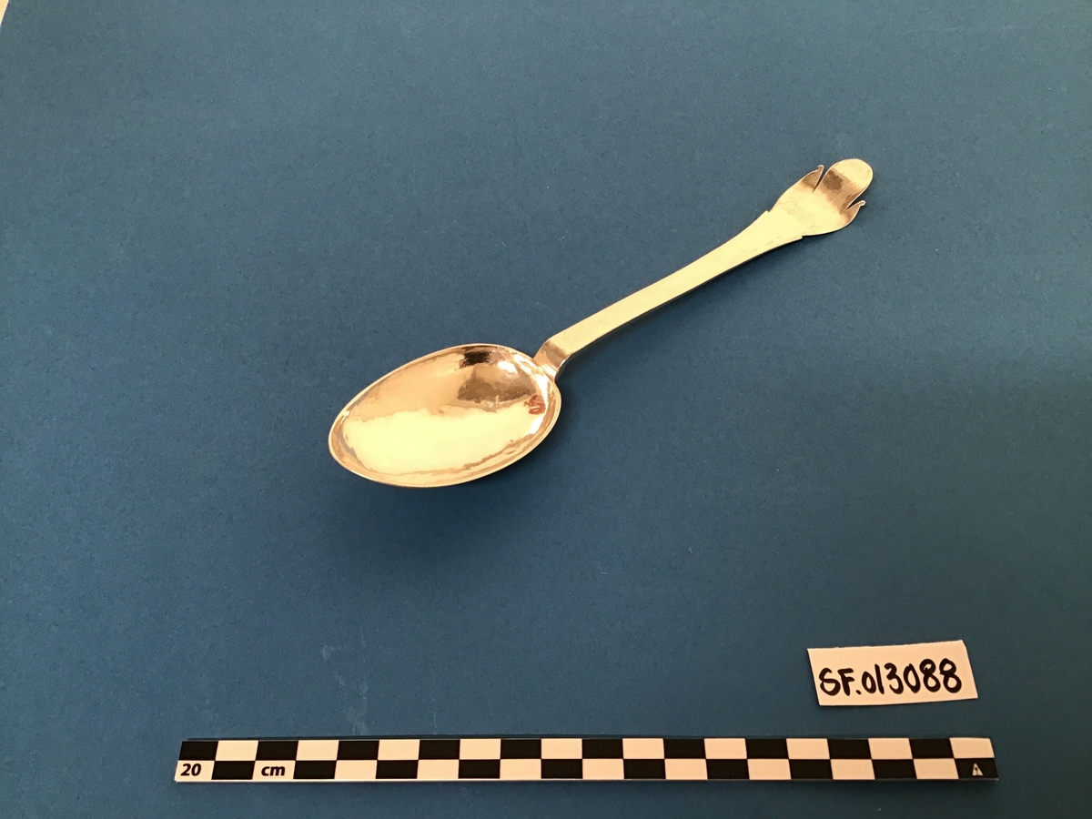 Sølvskei.Tilnærma elipseforma blad På baksida av blad innriss av eit ornament. Skaftet er bøygd opp i enden. På baksida her finn ein stempel og initialar.