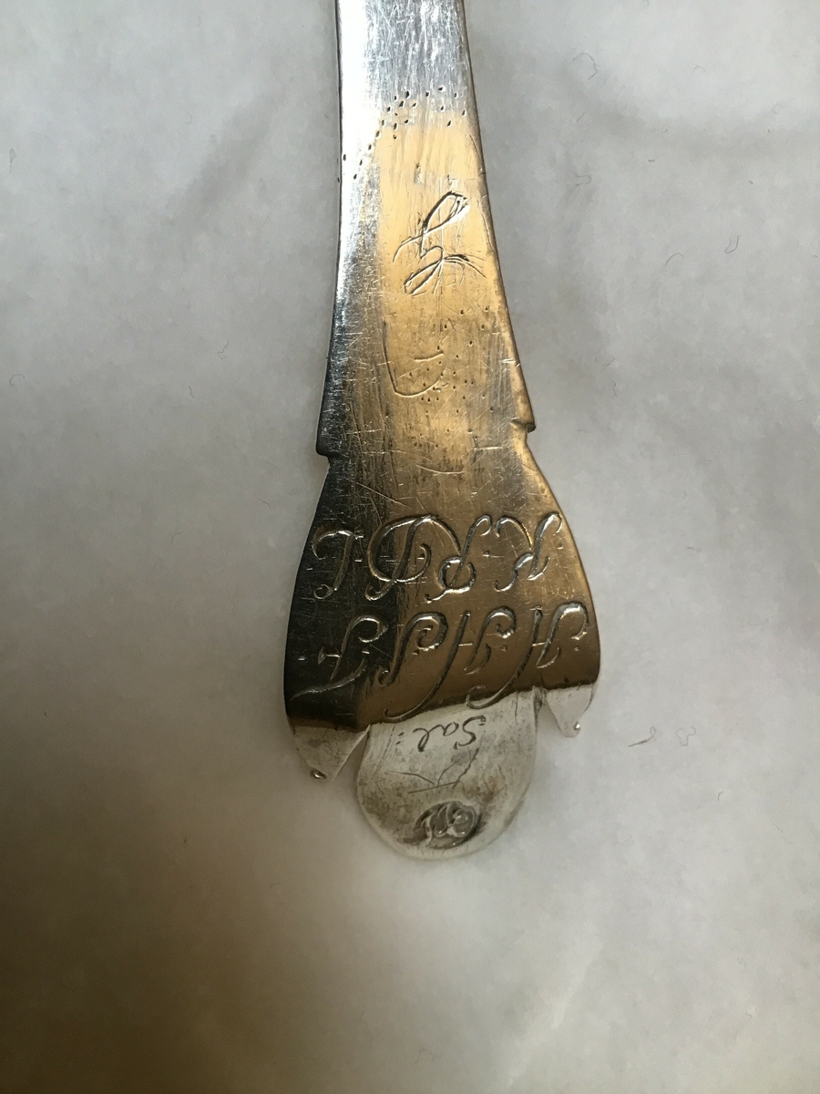 Sølvskei.Tilnærma elipseforma blad På baksida av blad innriss av eit ornament. Skaftet er bøygd opp i enden. På baksida her finn ein stempel og initialar.