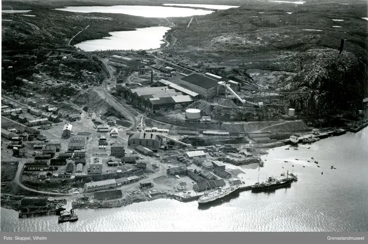Flyfoto over AS Sydvaranger med havn og andre anlegg, mot Første-, Andre- og Tredjevann. Deler av Kirkenes sentrum til venstre. Hurtigruta ligger til kai. Dampsentralen mot midten av bildet.