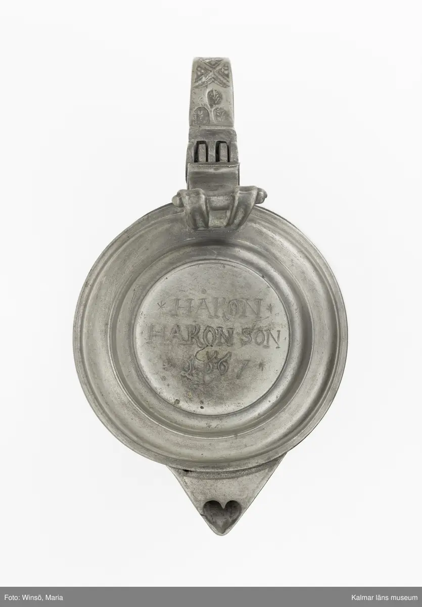 KLM 7267. Kanna, skänkkanna. Av tenn. Cylindrisk kanna med snås, lock och hänkel med tumgrepp. På locket inskription: Håkon Hakonson 1667. På grepen upptill 3 stämplar: Kalmar, två mästarstämplar, LS= Lars Sigfridsson. Använd för nattvardsvin men troligen från början för profant bruk.