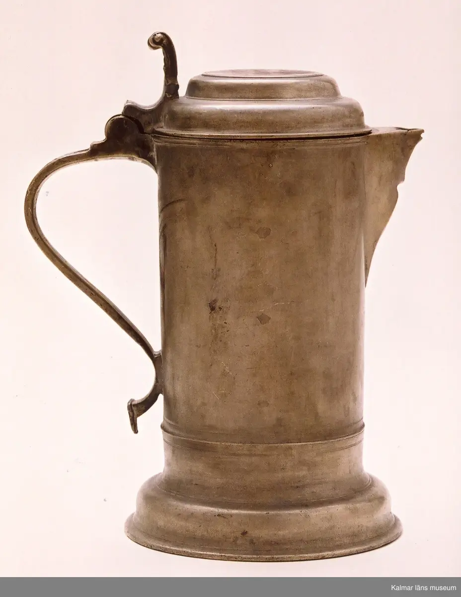KLM 7267. Kanna, skänkkanna. Av tenn. Cylindrisk kanna med snås, lock och hänkel med tumgrepp. På locket inskription: Håkon Hakonson 1667. På grepen upptill 3 stämplar: Kalmar, två mästarstämplar, LS= Lars Sigfridsson. Använd för nattvardsvin men troligen från början för profant bruk.