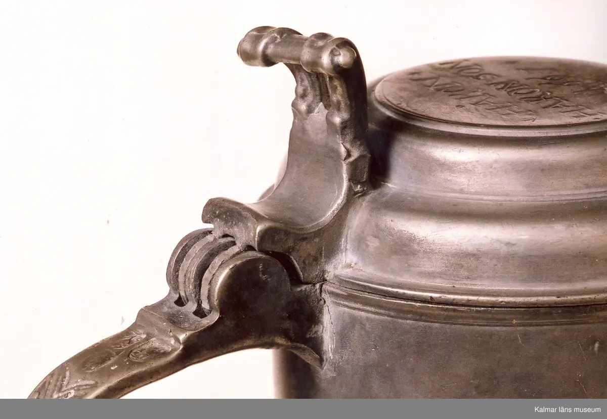 KLM 7267. Kanna, skänkkanna. Av tenn. Cylindrisk kanna med snås, lock och hänkel med tumgrepp. På locket inskription: Håkon Hakonson 1667. På grepen upptill 3 stämplar: Kalmar, två mästarstämplar, LS= Lars Sigfridsson. Använd för nattvardsvin men troligen från början för profant bruk.