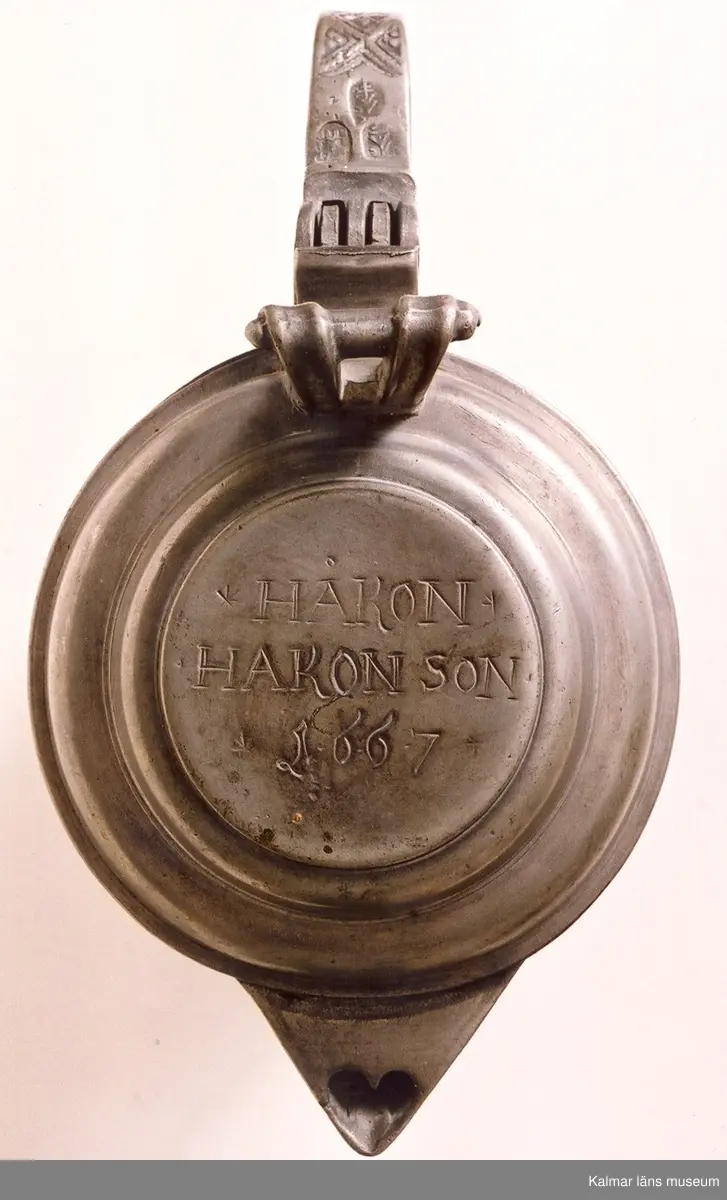 KLM 7267. Kanna, skänkkanna. Av tenn. Cylindrisk kanna med snås, lock och hänkel med tumgrepp. På locket inskription: Håkon Hakonson 1667. På grepen upptill 3 stämplar: Kalmar, två mästarstämplar, LS= Lars Sigfridsson. Använd för nattvardsvin men troligen från början för profant bruk.