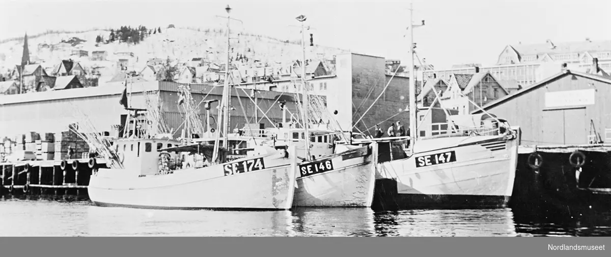 Danske laksefiskere i havn i Harstad, tre fiskebåter. Kaia utenfor Stål og Jern. Vi ser den nærmeste bebyggelsen omkring. Ei kirke til venstre på bildet.