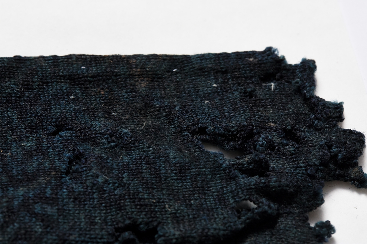 Strikk, kan det være strømpe? eller vott? Sist brukt som fyll i dør eller vinduskarm.
Prøven er glattstrikket med 5 masker pr cm. Rundstrikket. Garnet er mørkt blått og lysere blått, melert. Raknekanter på alle sider.
I ene enden er det sydd fast, med dobbel lintråd og attersting/kontursting, en tøybit, brettet til et slags bånd. Tøyet er brunt, toskaftsvevd av ull. "Båndet er slitt av. Lengde 4,5 cm, bredde 0,75-1 cm. Kan båndet ha vært en hempe?
b)funnet seinere. Rundstrikket. Brun tråd i ene enden. Stopping?