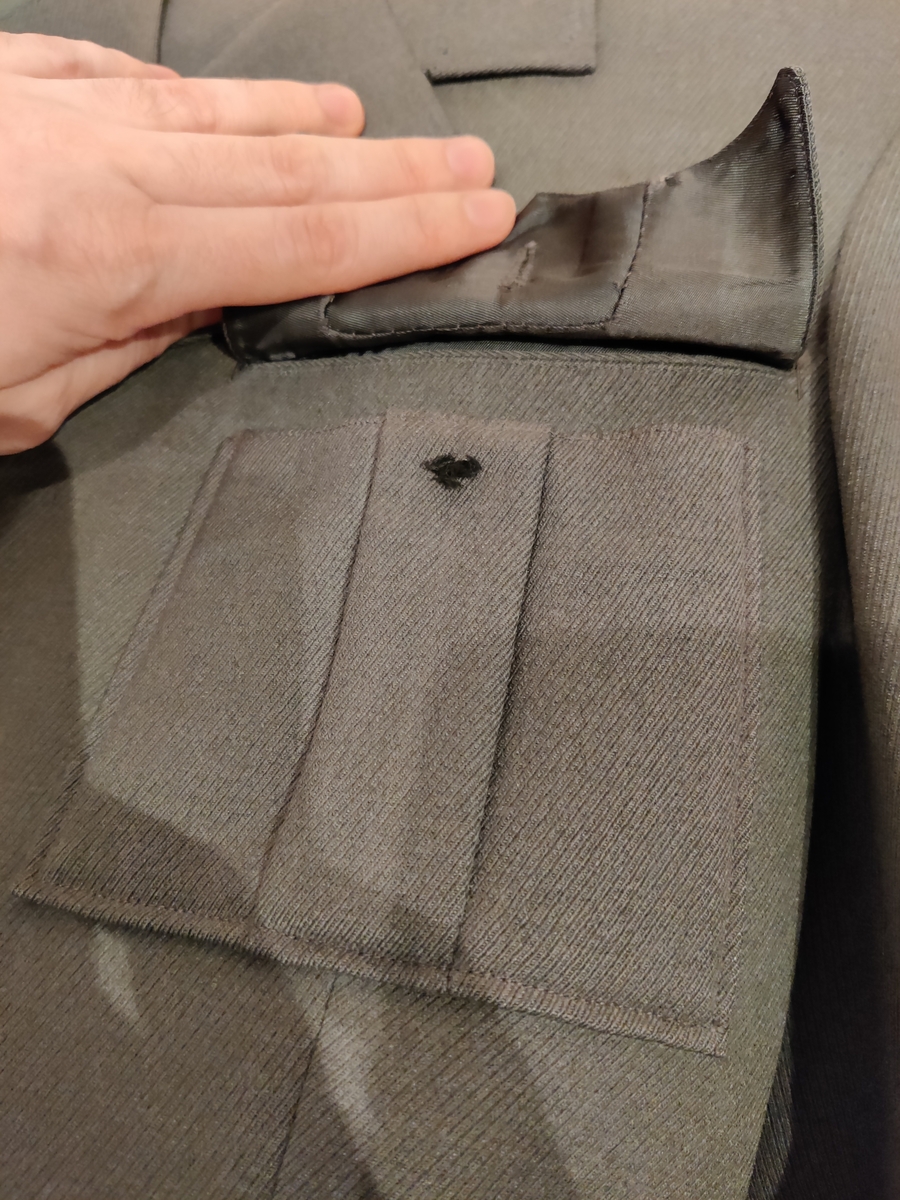Grågrønn uniform med liggende krave og fire lommer, ridebukse og skilue (Finn Quales modell). Uniformen har bruksmerker, særlig på kraven. Luen er slitt, foret har løsnet. Alle gradstegn og knapper er fjernet fra jakken. God kvalitet på stoffet, muligens offisersuniform. (NB: Fremstår som grå på bildene, men har i virkeligheten en blass militærgrønn farge.)