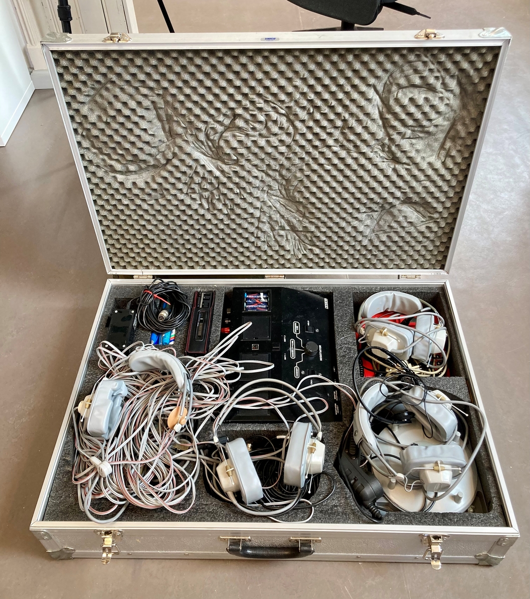 Koffert i metall som inneholder telefon og telefonsentral, opptaksutstyr og flere øretelefoner, lange telefonledninger, samt noe verktøy og kontorrekvisita, som skrujern, tapetkniv, tusjer, skrujern notatpapir, stifter, ekstra batterier mm.