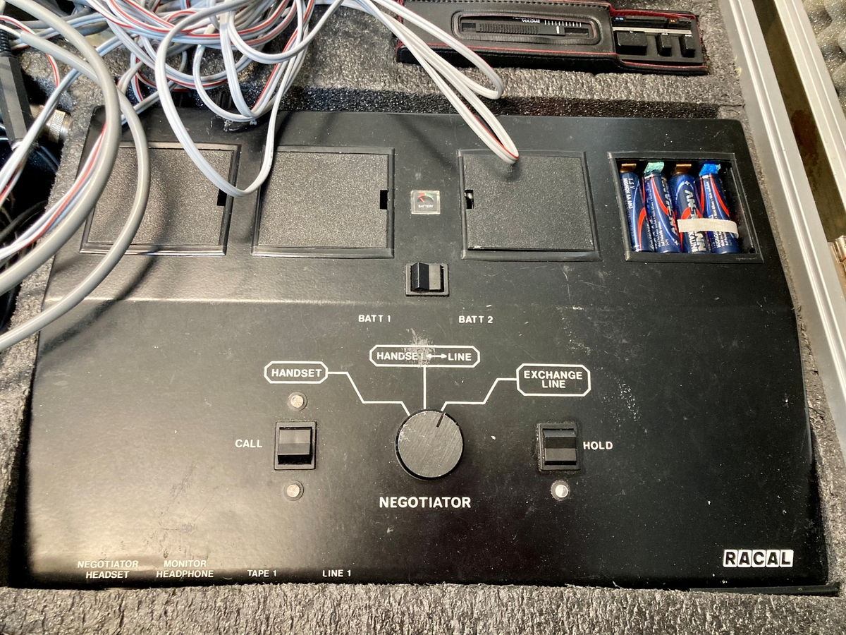 Koffert i metall som inneholder telefon og telefonsentral, opptaksutstyr og flere øretelefoner, lange telefonledninger, samt noe verktøy og kontorrekvisita, som skrujern, tapetkniv, tusjer, skrujern notatpapir, stifter, ekstra batterier mm.