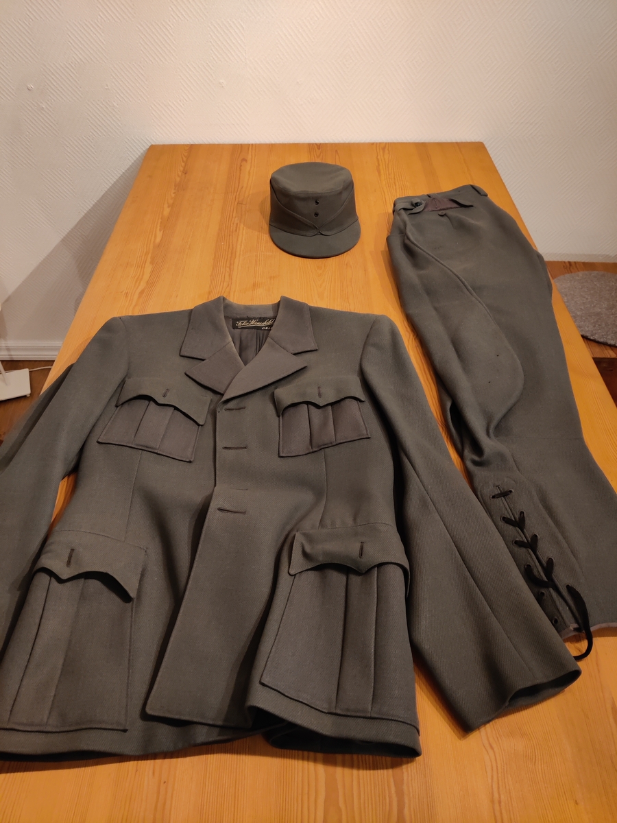 Grågrønn uniform med liggende krave og fire lommer, ridebukse og skilue (Finn Quales modell). Uniformen har bruksmerker, særlig på kraven. Luen er slitt, foret har løsnet. Alle gradstegn og knapper er fjernet fra jakken. God kvalitet på stoffet, muligens offisersuniform. (NB: Fremstår som grå på bildene, men har i virkeligheten en blass militærgrønn farge.)