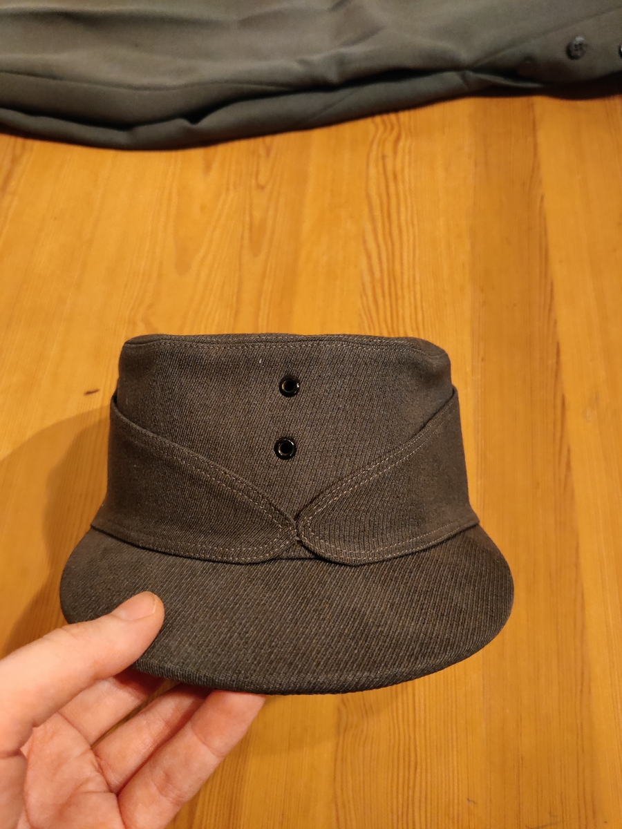 Grågrønn uniform med liggende krave og fire lommer, ridebukse og skilue (Finn Quales modell). Uniformen har bruksmerker, særlig på kraven. Luen er slitt, foret har løsnet. Alle gradstegn og knapper er fjernet fra jakken. God kvalitet på stoffet, muligens offisersuniform. (NB: Fremstår som grå på bildene, men har i virkeligheten en blass militærgrønn farge.)