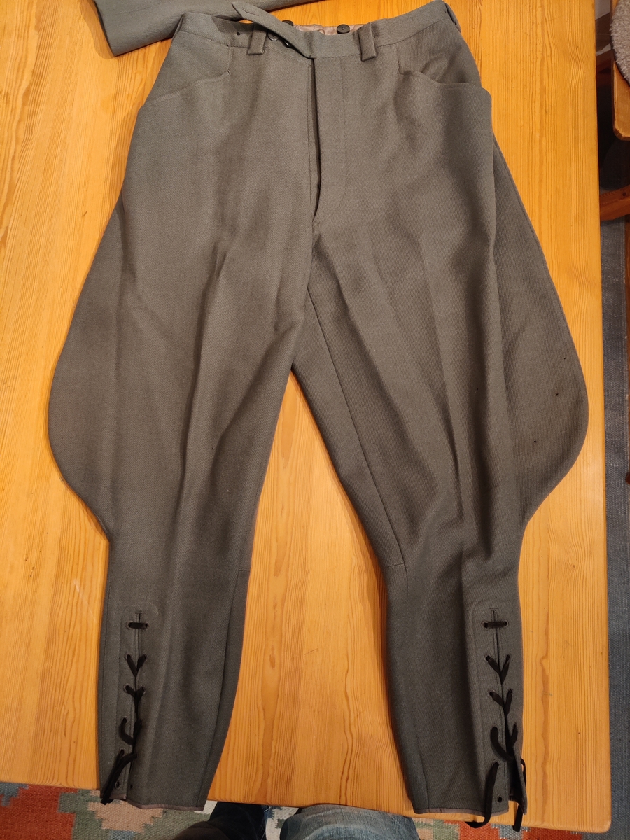 Grågrønn uniform med liggende krave og fire lommer, ridebukse og skilue (Finn Quales modell). Uniformen har bruksmerker, særlig på kraven. Luen er slitt, foret har løsnet. Alle gradstegn og knapper er fjernet fra jakken. God kvalitet på stoffet, muligens offisersuniform. (NB: Fremstår som grå på bildene, men har i virkeligheten en blass militærgrønn farge.)