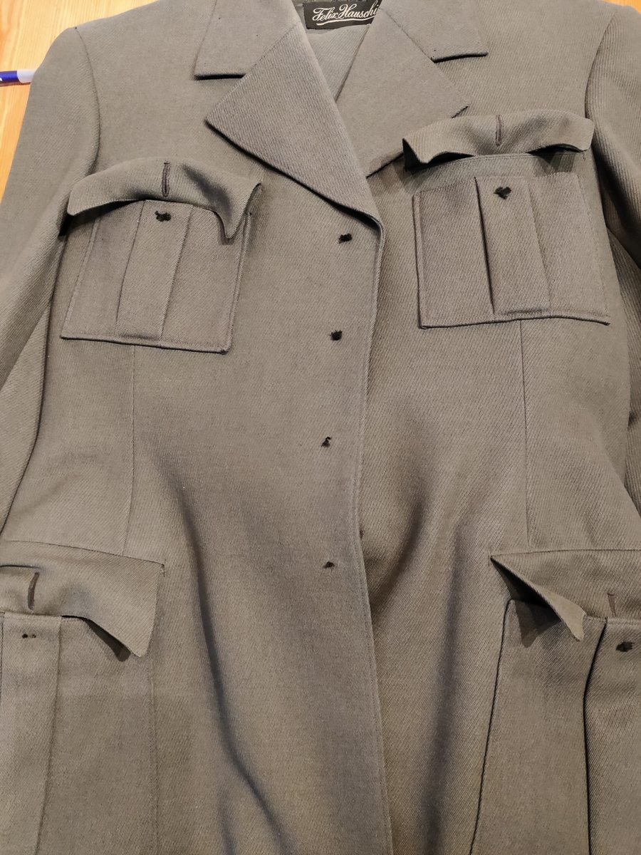 Grågrønn uniform med liggende krave og fire lommer, ridebukse og skilue (Finn Quales modell). Uniformen har bruksmerker, særlig på kraven. Luen er slitt, foret har løsnet. Alle gradstegn og knapper er fjernet fra jakken. God kvalitet på stoffet, muligens offisersuniform. (NB: Fremstår som grå på bildene, men har i virkeligheten en blass militærgrønn farge.)