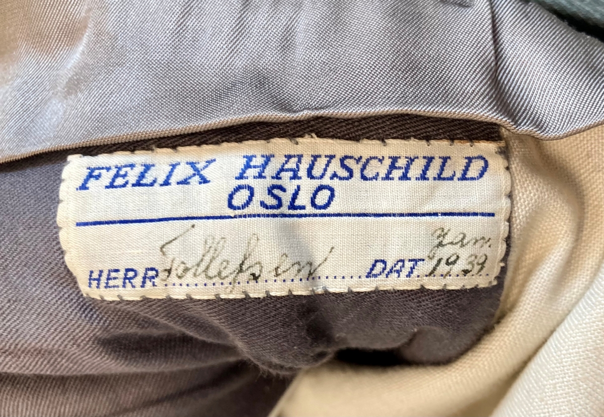 Grågrønn uniform med liggende krave og fire lommer, ridebukse og skilue (Finn Quales modell). Uniformen har bruksmerker, særlig på kraven. Luen er slitt, foret har løsnet. Alle gradstegn og knapper er fjernet fra jakken. God kvalitet på stoffet, muligens offisersuniform. (NB: Fremstår som grå på bildene, men har i virkeligheten en blass militærgrønn farge.)