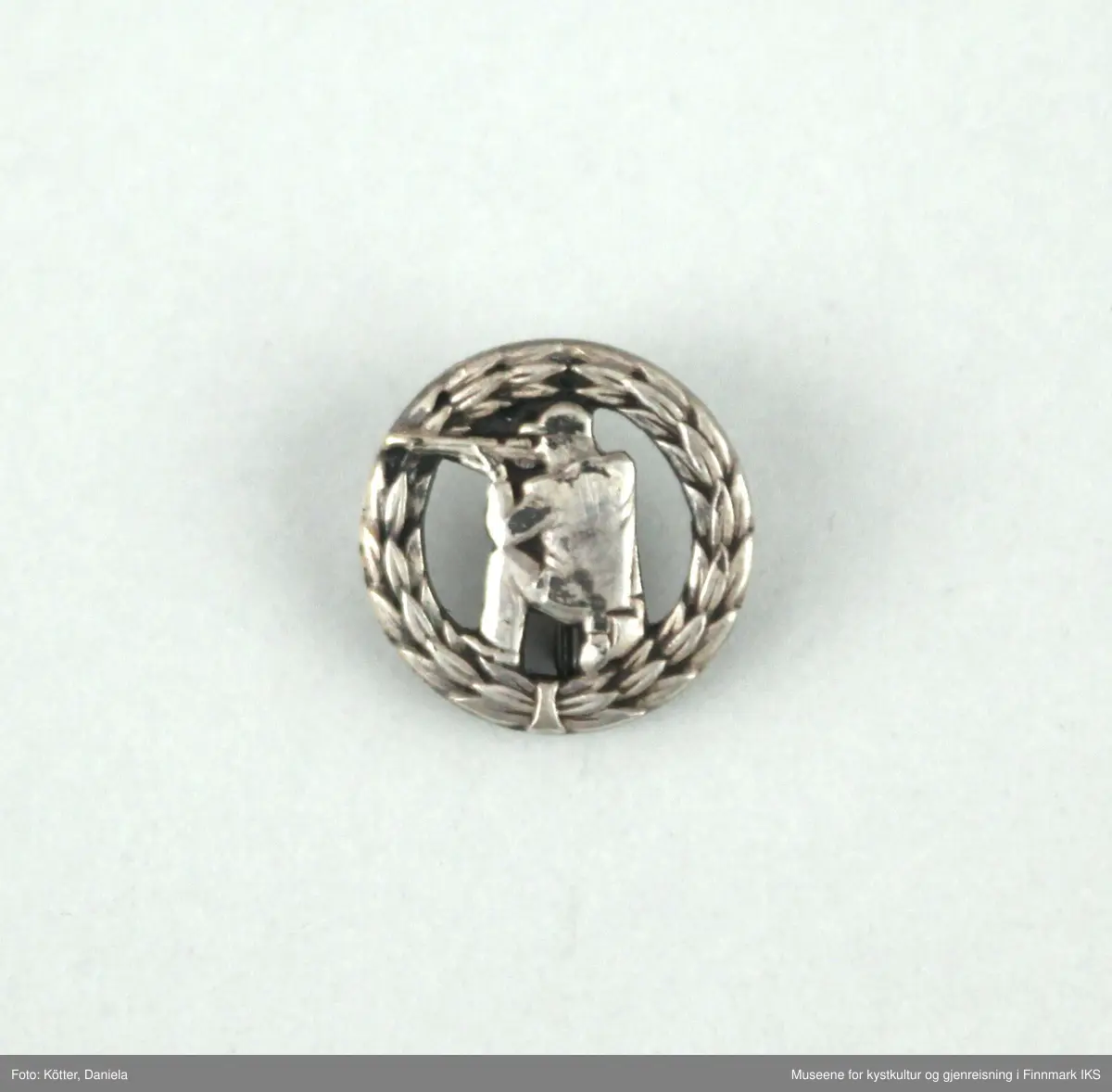 Merket er i sølv og fremstiller en rund laurbærkrans som omfavner en knestående skytter som sikter. Merket festes med et nål som er påført knappens bakside.