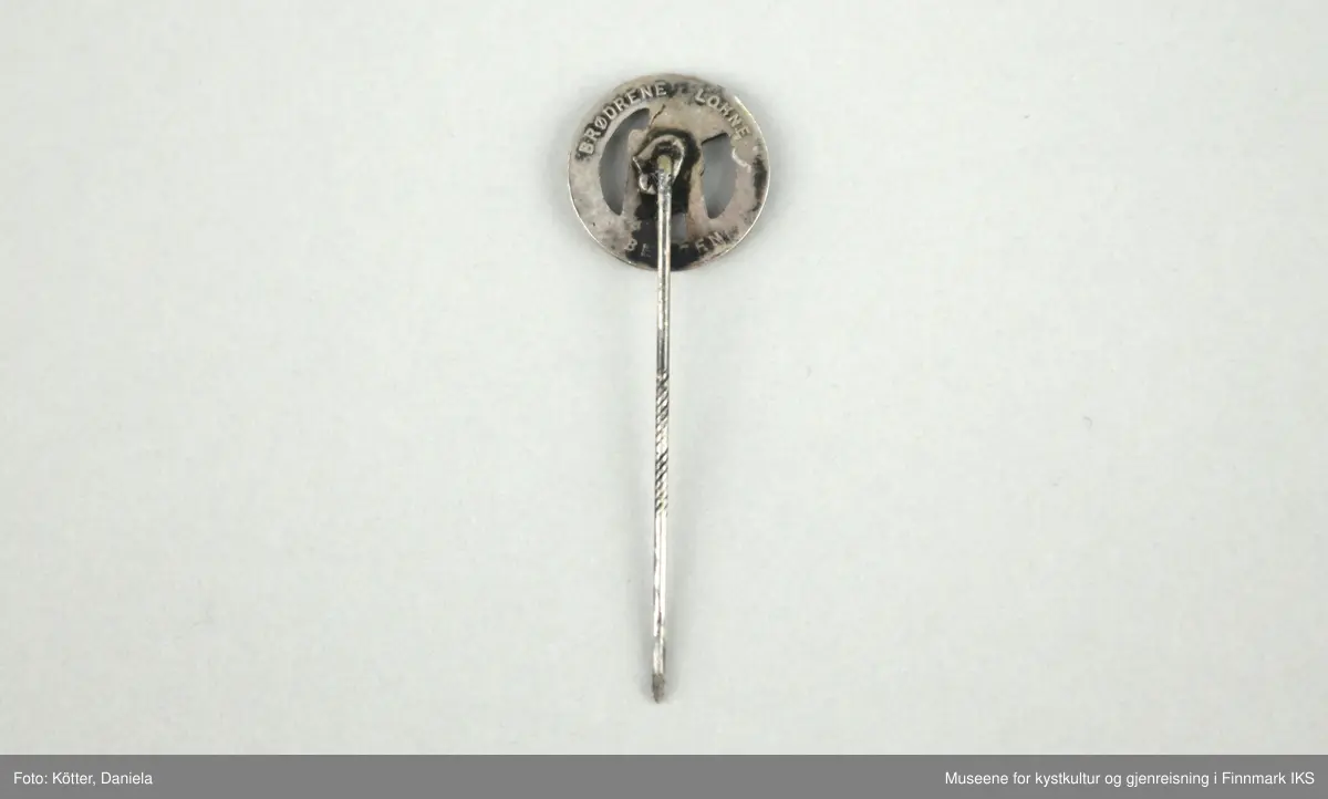 Merket er i sølv og fremstiller en rund laurbærkrans som omfavner en knestående skytter som sikter. Merket festes med et nål som er påført knappens bakside.