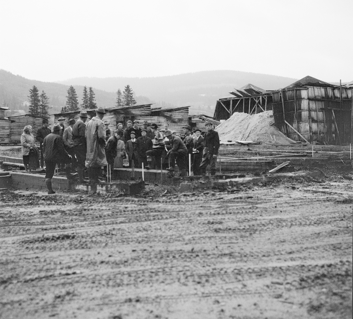 Fra sagtomta ved Trysil kommunale sag og høvleri. Dette fotografiet er antakelig tatt under ei befaring Trysil kommunale skogforvaltning arrangerte for lokalpolitikerne høsten 1964, da virksomheten var i en ekspansjonsfase med tunge investeringer som forutsatte politikernes aksept. Disponent Jostein Bjørnersen gjorde sitt beste for å overbevise representantene for eierskapet om at dette var et godt prosjekt, som ville føre til at tryslingene fikk høste de økonomiske gevinstene det innebar å foredle tømmer fra lokale skoger, i første instans til trelast, og deretter til bygging av hytter og hus. Denne virksomheten skulle skape arbeidsplasser og bidra til velstand i bygda. Da dette fotografiet ble tatt var politkerne samlet for å høre disponentens orientering på sagtomta. I bakgrunnen ser vi bordstabler som ble tørket i friluft og avfallsflis fra det eksisterende sagbruket.