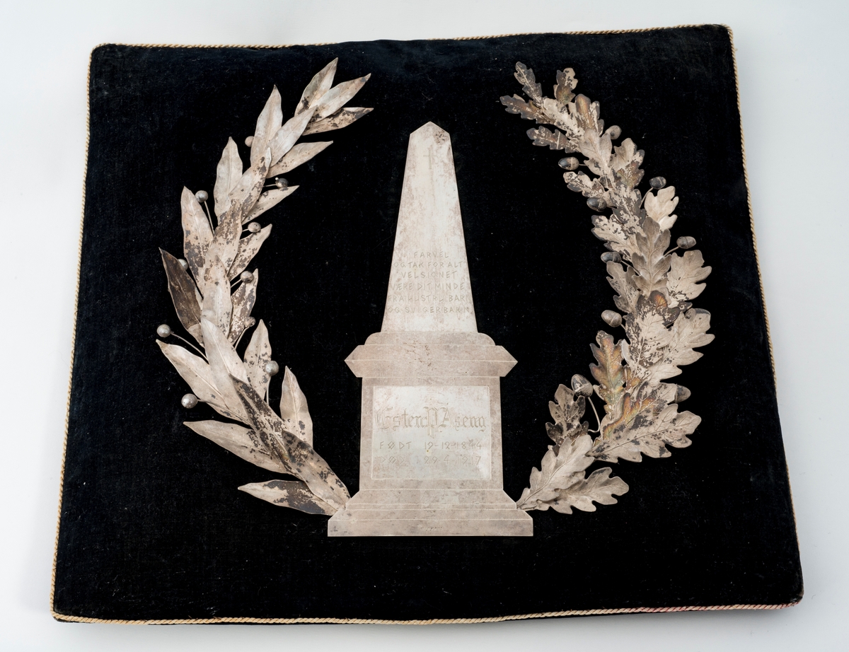 Krans på pute. Den ene delen av kransen har eikeblad og eikenøtter som motiv, og den andre delen har en flatere type blad med knopper. I midten er det en obelisk på en plattform.