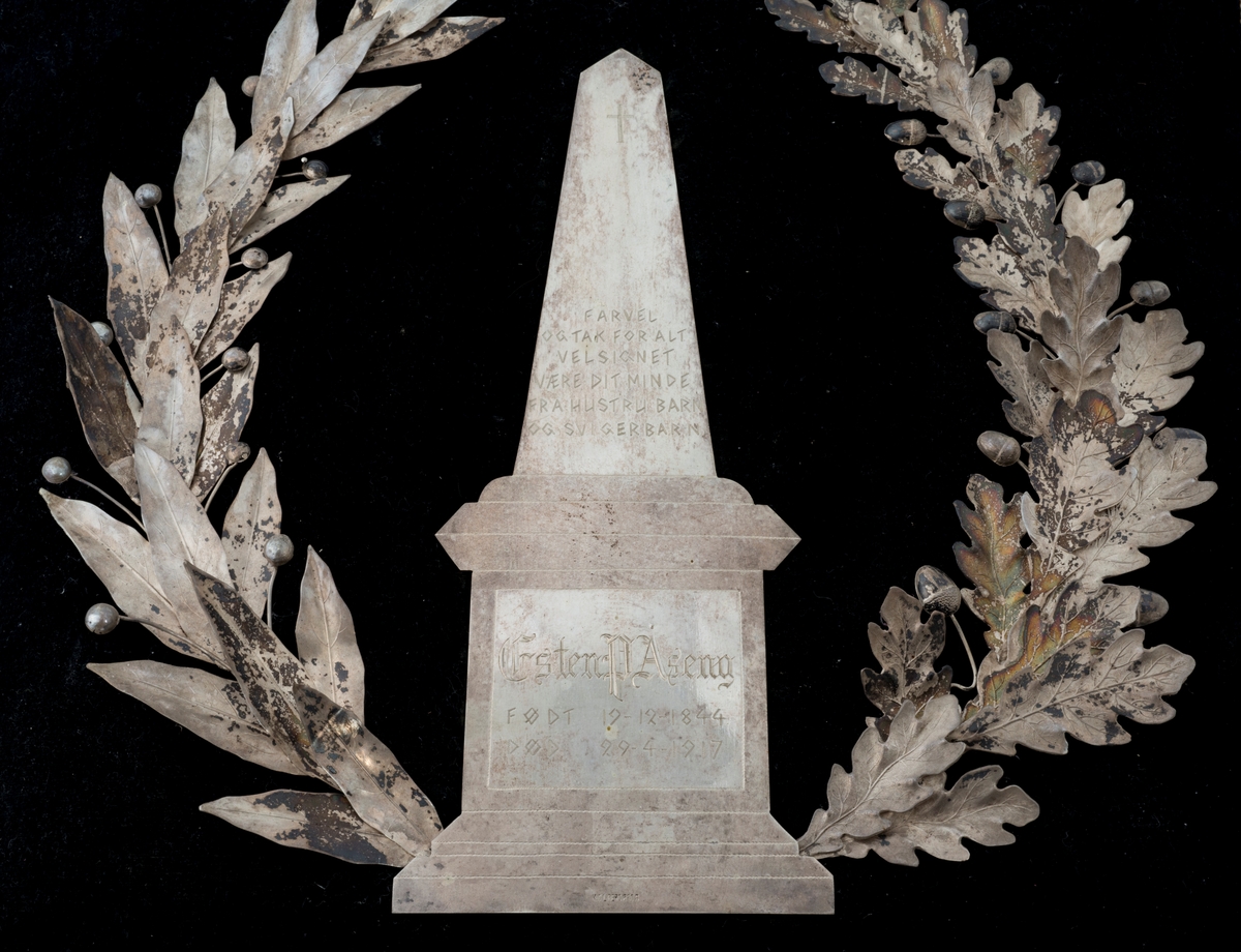 Krans på pute. Den ene delen av kransen har eikeblad og eikenøtter som motiv, og den andre delen har en flatere type blad med knopper. I midten er det en obelisk på en plattform.
