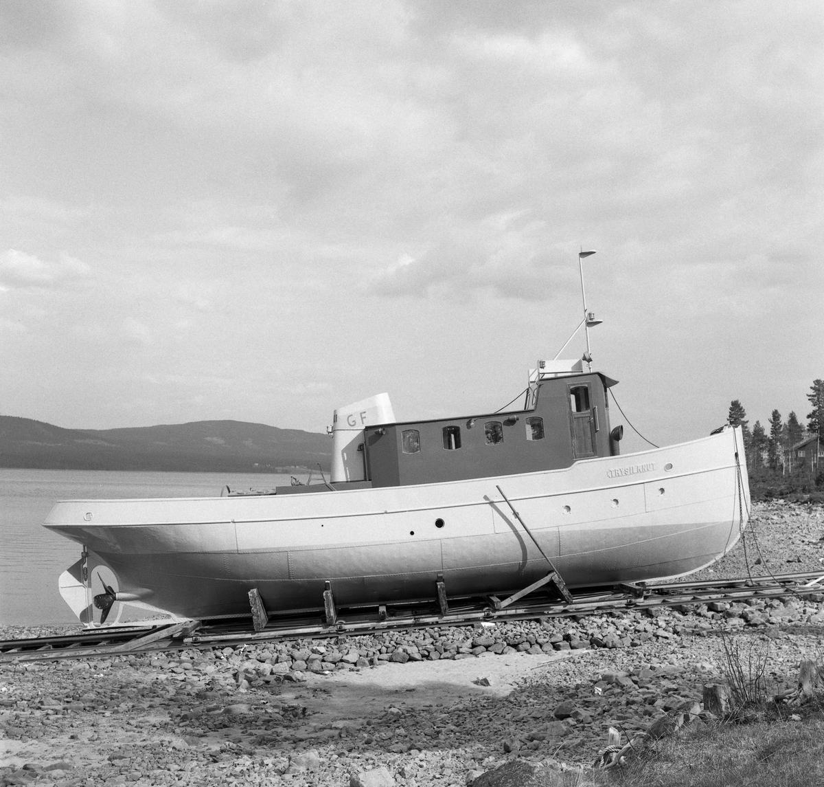 Slepebåten «Trysilknut», fotografert ved slippen på Sørlistøa ved Osensjøen i Åmot kommune i Hedmark i 1964. Fartøyet er fotografert fra styrbord side. I 1964 var dette fartøyet 50 år gammelt. Skroget var slik det alltid hadde vært - 55 fot langt og 12 fot bredt, klinket sammen av det som i 1914 var «bedste sort skibsbygningsstaal», levert av Glommens mek. Verksted i Fredrikstad. Men Trysilknut var i utgangspunktet et dampdrevet fartøy. Dampmaskinen viste seg imidlertid i veikeste laget for de store tømmerbommene som skulle bukseres over den bortimot tre mil lange og 47 kvadratkilometer store Osensjøen. Etter 2. verdenskrig ble mange av slepebåtene som fellesfløtingsforeningene disponerte ombygd til dieseldrift. Ved å gjøre en slik investering så man for seg at man på litt lengre sikt skulle oppnå større trekkraft og samtidig redusere driftskostandene. Dieselmotorene krevde nemlig langt mindre betjening enn dampmaskinene, og følgelig ville en dieseldrevet slepebåt kunne opereres med mindre mannskap enn et dampfartøy. I Trysilknuts tilfelle så man for seg at to fyrbøtere og en maskinist kunne utgå fra besetningen. Her lå det et betydelig innsparingspotensial i en periode med stigende lønnskostnader. I 1956 sendte direktøren i Glomma fellesfløtingsforening prisforespørsler til firmaer som man antok kunne påta seg ombyggingsarbeidet. Oppdraget gikk også denne gangen til Glommens mek. verksted. Demonteringa av dampmaskinen innebar også at det ble en del ledig plass om bord. Ominnredningsarbeidet ble overlatt til Ivar Hagens båtbyggeri på Hjelset utenfor Molde. Resultatet ble at Trysilknut - etter ombyggingsarbeidet vinteren 1957-58 - framsto med et noe endret eksteriør og et fullstendig modernisert interiør.