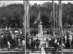 Fotografi i samband med invigningen av Per Brahe-bysten på Gränna torg 1916. En folksamling kring bysten och en talarstol med en man till vänster om densamma. Flaggor kantar vägen från Brahegatan upp mot bysten.