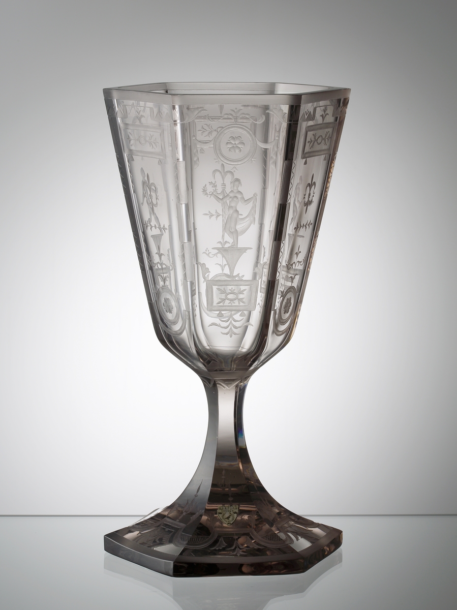Design: Simon Gate. Graverad av Emil Weidlich (Orrefors 1922-29). Pokal med sexkantig kupa, sexkantigt ben och fot. Ytorna är plan- och facettslipade, och försedda med gravyrer bestående av baldakiner, växtmotiv och oklädda män och kvinnor.
Etikett: Vit botten, svart text och orre.