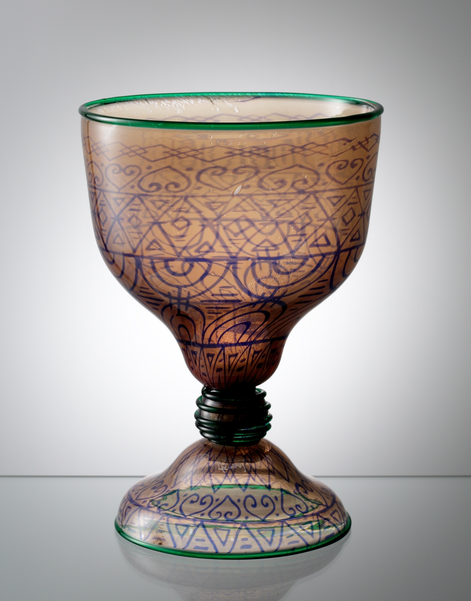 Graalskål nr 122, formgiven av Simon Gate, mönstermakare Heinrich Wollman, mästerblåsare Knut Bergqvist. Blått geometriskt mönster mot en rosatonad botten. På skålens mynning och fotens kant sitter påklippta gröna trådar, samt en spunnen grön tråd på kulan mellan skål och fot.