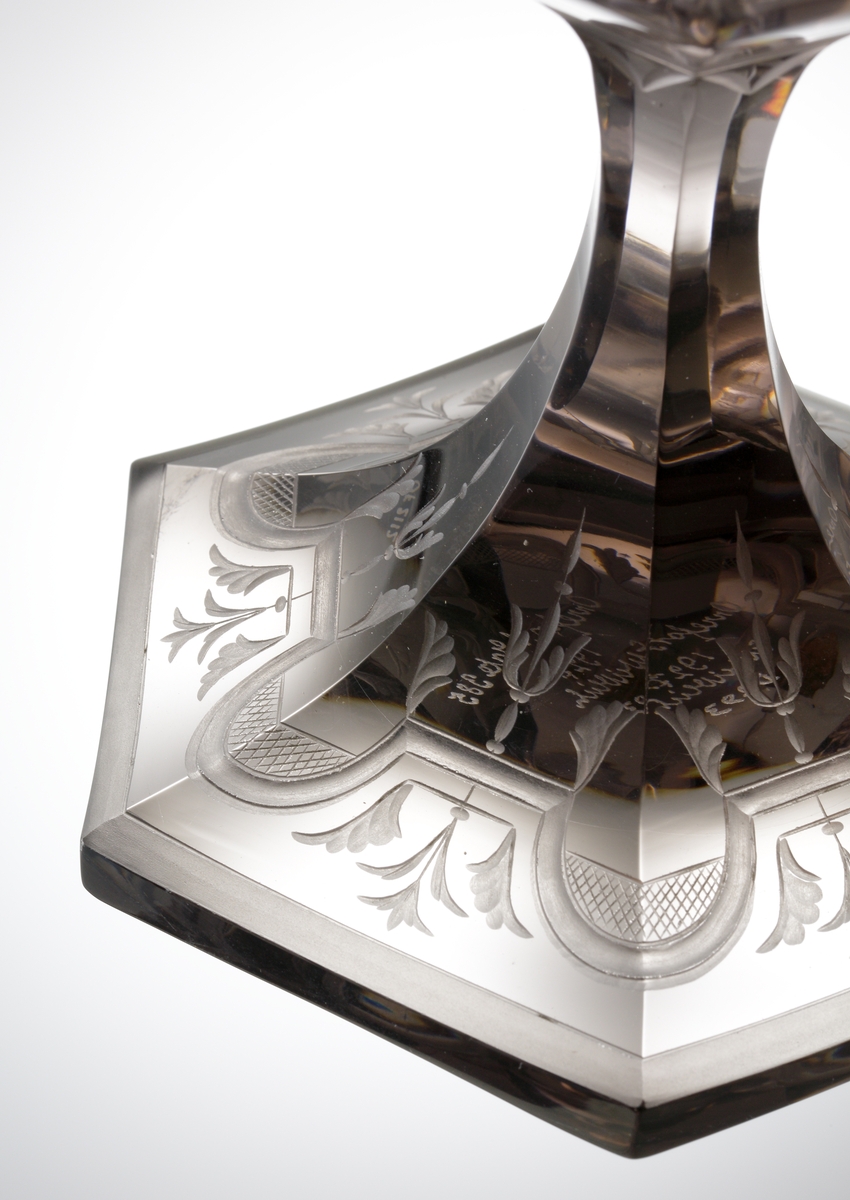 Design: Simon Gate. Graverad av Emil Weidlich (Orrefors 1922-29). Pokal med sexkantig kupa, sexkantigt ben och fot. Ytorna är plan- och facettslipade, och försedda med gravyrer bestående av baldakiner, växtmotiv och oklädda män och kvinnor.
Etikett: Vit botten, svart text och orre.