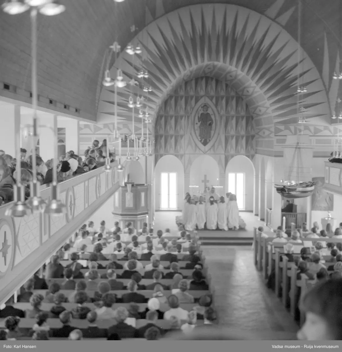 Vadsø kirke, interiør 10.5.1969. Konfirmasjon, konfirmanter kneler ved alteret. Kirka ble tegnet av arkitekt Magnus Poulsson og vigslet i 1958.