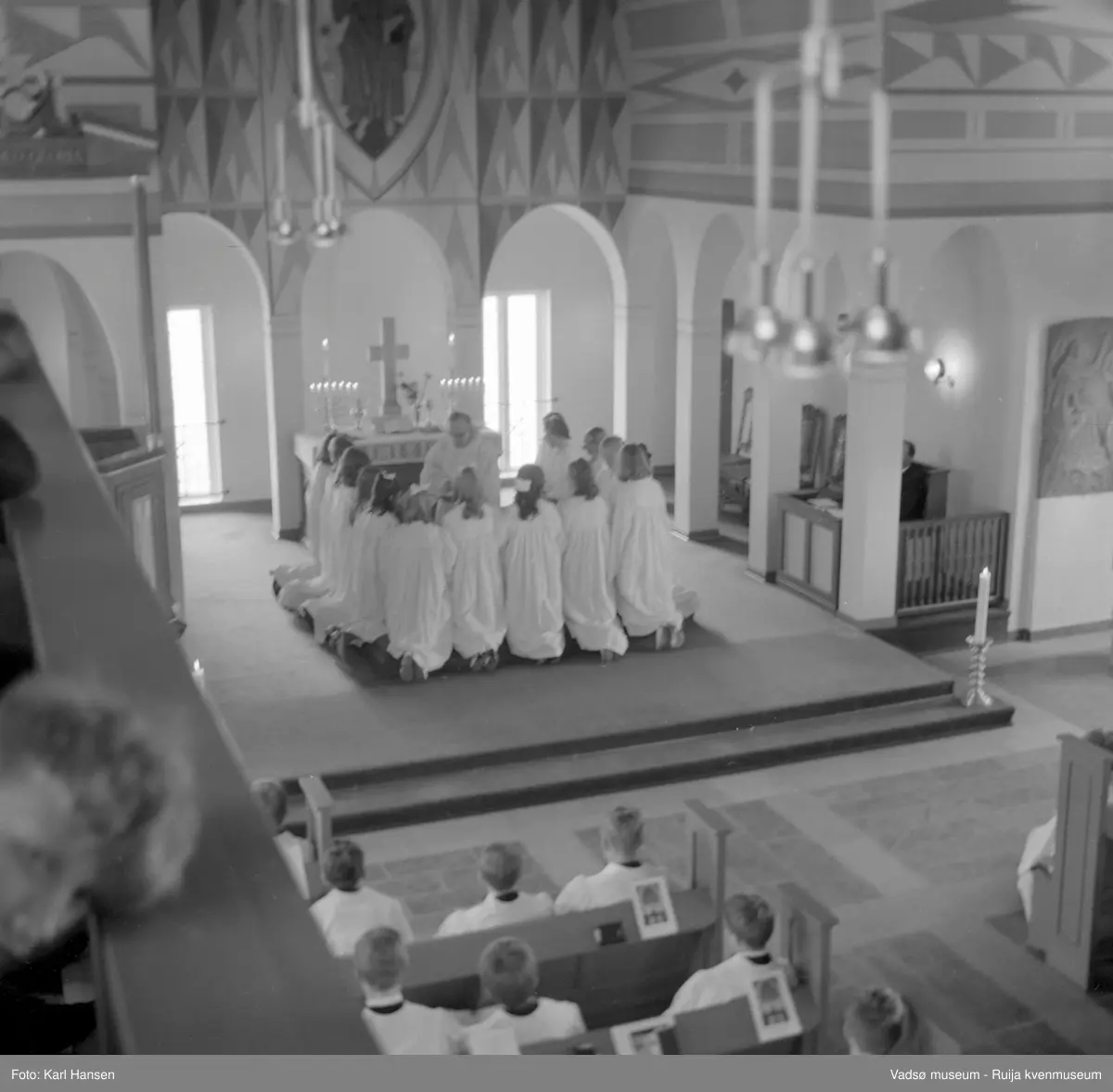 Vadsø kirke, interiør 10.5.1969. Konfirmasjon, konfirmanter kneler ved alteret. Kirka ble tegnet av arkitekt Magnus Poulsson, vigslet i 1958.