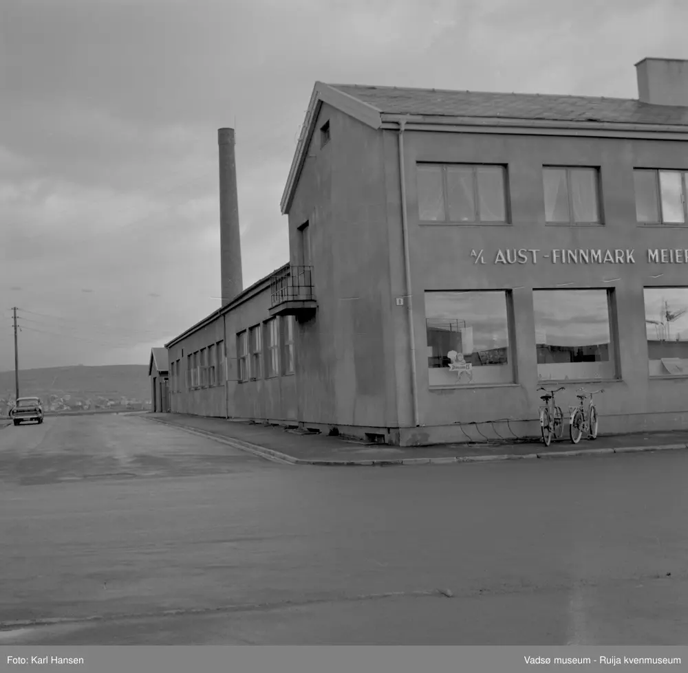 A/L Aust-Finnmark Meieri, 1962 i Vadsø. Bildet er tatt mot øst og krysset av Tollbugata og W. Andersensgate.
Arbeidet med å få meieri til Vadsø ble påbegynt før krigen.  Ønsket var å få et meieri som kunne dekke Nord-Varanger-Tanadistriktet.  Andelslaget A/L Aust-Finnmark Meieri ble stiftet 28.juni 1947 i Nyborg.  
Aust- Finnmark Meieri ble oppført i Tollbugata i 1949; arkitekt Monrad Heggen m hovedentreprenør Brdr. Aronsen.
Oppstart skulle finne sted ca 1. oktober 1949, og i mai ble det ansatt en meieriarbeider; Richard Korbi fra Vadsø.
Kilde: "A.L Aust-Finnmark Meieri 25 års jubileum 1949-1974; styreformann Ivar Johnsen, Tana mai 1974"