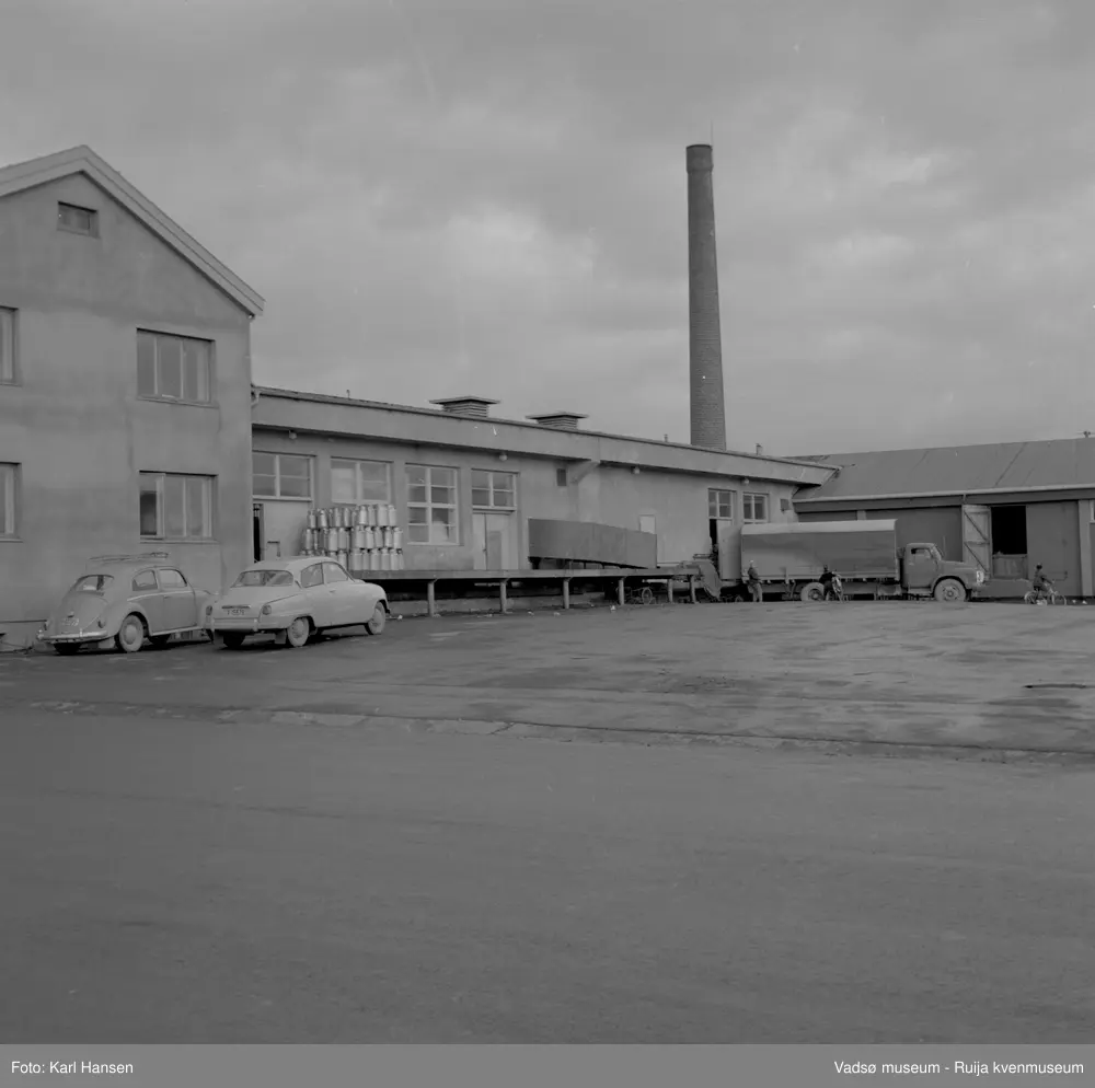 A/L Aust-Finnmark Meieri, 1962 i Vadsø. Bildet er tatt mot nordøst. En lastebil er rygget inn til bygningen for av/pålasting og  ved bilen to-tre personer med sykkel. På lasterampen står stabler av melkespann og to personbiler ved husveggen. 
Arbeidet med å få meieri til Vadsø ble påbegynt før krigen.  Ønsket var å få et meieri som kunne dekke Nord-Varanger-Tanadistriktet.  Andelslaget A/L Aust-Finnmark Meieri ble stiftet 28.juni 1947 i Nyborg.  
Aust- Finnmark Meieri ble oppført i Tollbugata i 1949; arkitekt Monrad Heggen m hovedentreprenør Brdr. Aronsen.
Oppstart skulle finne sted ca 1. oktober 1949, og i mai ble det ansatt en meieriarbeider; Richard Korbi fra Vadsø.
Kilde: "A.L Aust-Finnmark Meieri 25 års jubileum 1949-1974; styreformann Ivar Johnsen, Tana mai 1974"
