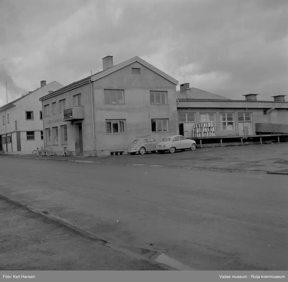 Aust-Finnmark Meieri, 1962 i Vadsø. Bildet er tatt mot nord. På lasterampen står stabler av melkespann og to personbiler ved husveggen. På fortauet står noen sykler parkert. Til venstre i bildet ser vi hjørnet av H.F. Esbensens forretningsbygg.
Arbeidet med å få meieri til Vadsø ble påbegynt før krigen.  Ønsket var å få et meieri som kunne dekke Nord-Varanger-Tanadistriktet.  Andelslaget A/L Aust-Finnmark Meieri ble stiftet 28.juni 1947 i Nyborg.  
Aust- Finnmark Meieri ble oppført i Tollbugata i 1949; arkitekt Monrad Heggen m hovedentreprenør Brdr. Aronsen.
Oppstart skulle finne sted ca 1. oktober 1949, og i mai ble det ansatt en meieriarbeider; Richard Korbi fra Vadsø.
Kilde: "A.L Aust-Finnmark Meieri 25 års jubileum 1949-1974; styreformann Ivar Johnsen, Tana mai 1974"