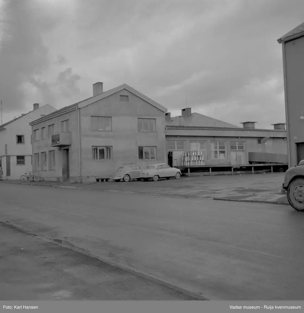 A/L Aust-Finnmark Meieri, 1962 i Vadsø. Bildet er tatt mot nord. På lasterampen står stabler av melkespann og to personbiler ved husveggen. På fortauet står noen sykler parkert. Til venstre i bildet ser vi hjørnet av H.F. Esbensens forretningsbygg.
Arbeidet med å få meieri til Vadsø ble påbegynt før krigen.  Ønsket var å få et meieri som kunne dekke Nord-Varanger-Tanadistriktet.  Andelslaget A/L Aust-Finnmark Meieri ble stiftet 28.juni 1947 i Nyborg.  
Aust- Finnmark Meieri ble oppført i Tollbugata i 1949; arkitekt Monrad Heggen m hovedentreprenør Brdr. Aronsen.
Oppstart skulle finne sted ca 1. oktober 1949, og i mai ble det ansatt en meieriarbeider; Richard Korbi fra Vadsø.
Kilde: "A.L Aust-Finnmark Meieri 25 års jubileum 1949-1974; styreformann Ivar Johnsen, Tana mai 1974"
