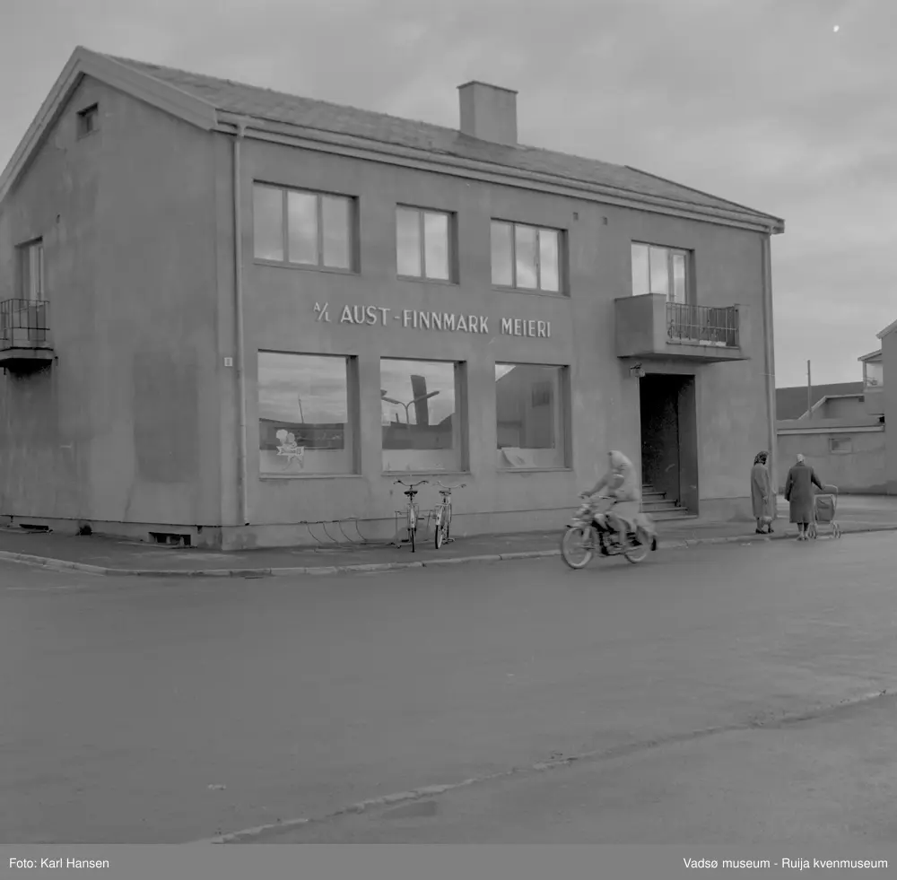 A/L Aust-Finnmark Meieri, 1962 i Vadsø. Bildet er tatt mot øst med oversikt over inngangspartiet og navnet A/L Aust-Finnmark på fasaden. Vi ser litt av W.A.Andersensgate til venstre i bildet.  På fortauet står noen sykler parkert og to damer som prater sammen. I Tollbugata kjører en dame forbi på moped. 
Arbeidet med å få meieri til Vadsø ble påbegynt før krigen.  Ønsket var å få et meieri som kunne dekke Nord-Varanger-Tanadistriktet.  Andelslaget A/L Aust-Finnmark Meieri ble stiftet 28.juni 1947 i Nyborg.  
Aust- Finnmark Meieri ble oppført i Tollbugata i 1949; arkitekt Monrad Heggen m hovedentreprenør Brdr. Aronsen.
Oppstart skulle finne sted ca 1. oktober 1949, og i mai ble det ansatt en meieriarbeider; Richard Korbi fra Vadsø.
Kilde: "A.L Aust-Finnmark Meieri 25 års jubileum 1949-1974; styreformann Ivar Johnsen, Tana mai 1974"