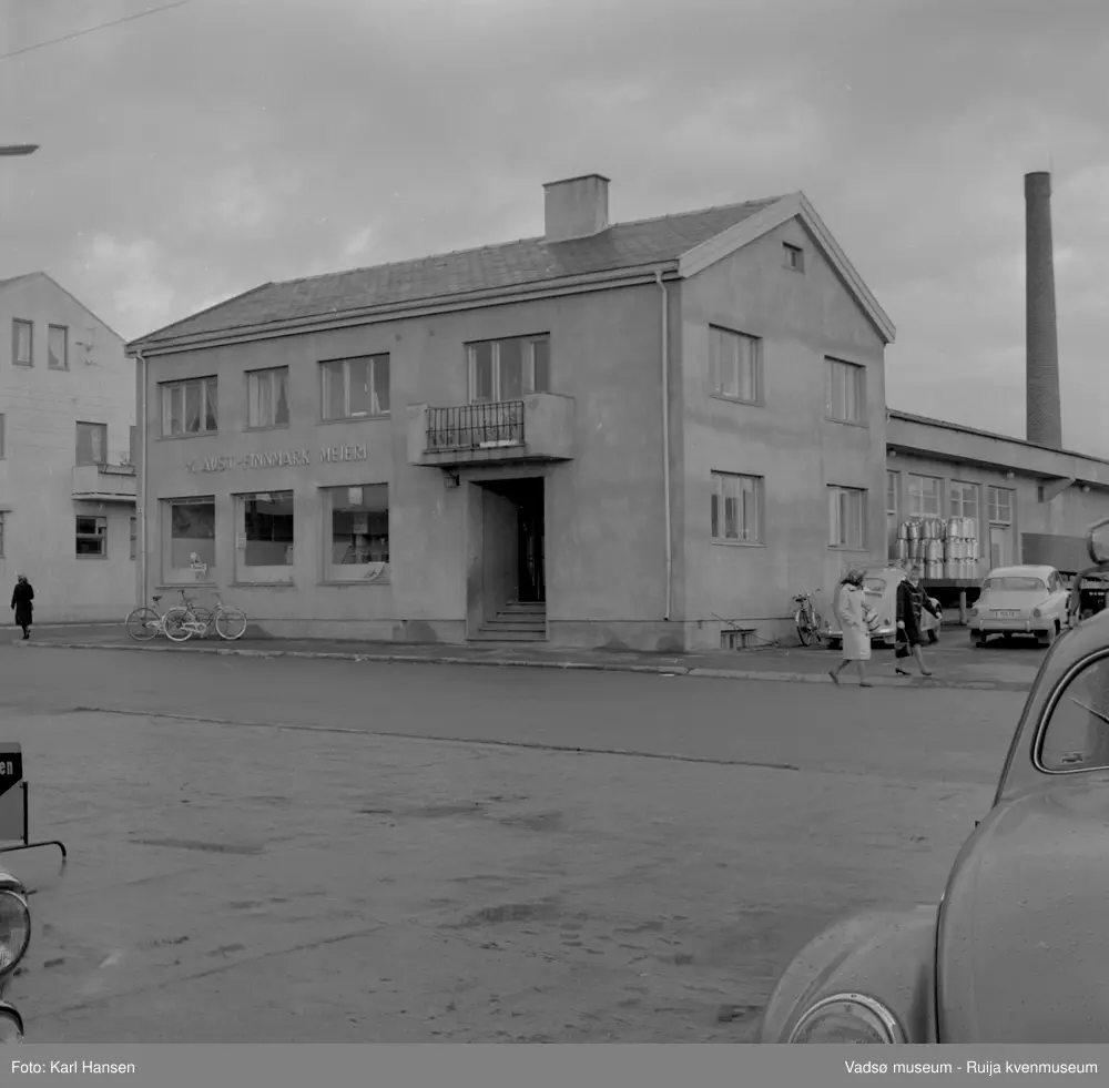 A/L Aust-Finnmark Meieri, 1962 i Vadsø. Bildet er tatt mot nordøst. Bilder viser inngangspartiet med fasaden samt lasterampen med stabler av melkespann. To biler og noen sykler står ved bygningen og vi ser også et par damet som spaserer på fortauet til Tollbugata. Til venstre i bildet ser vi hjørnet av H.F. Esbensens forretningsbygg.
Arbeidet med å få meieri til Vadsø ble påbegynt før krigen.  Ønsket var å få et meieri som kunne dekke Nord-Varanger-Tanadistriktet.  Andelslaget A/L Aust-Finnmark Meieri ble stiftet 28.juni 1947 i Nyborg.  
Aust- Finnmark Meieri ble oppført i Tollbugata i 1949; arkitekt Monrad Heggen m hovedentreprenør Brdr. Aronsen.
Oppstart skulle finne sted ca 1. oktober 1949, og i mai ble det ansatt en meieriarbeider; Richard Korbi fra Vadsø.
Kilde: "A.L Aust-Finnmark Meieri 25 års jubileum 1949-1974; styreformann Ivar Johnsen, Tana mai 1974"