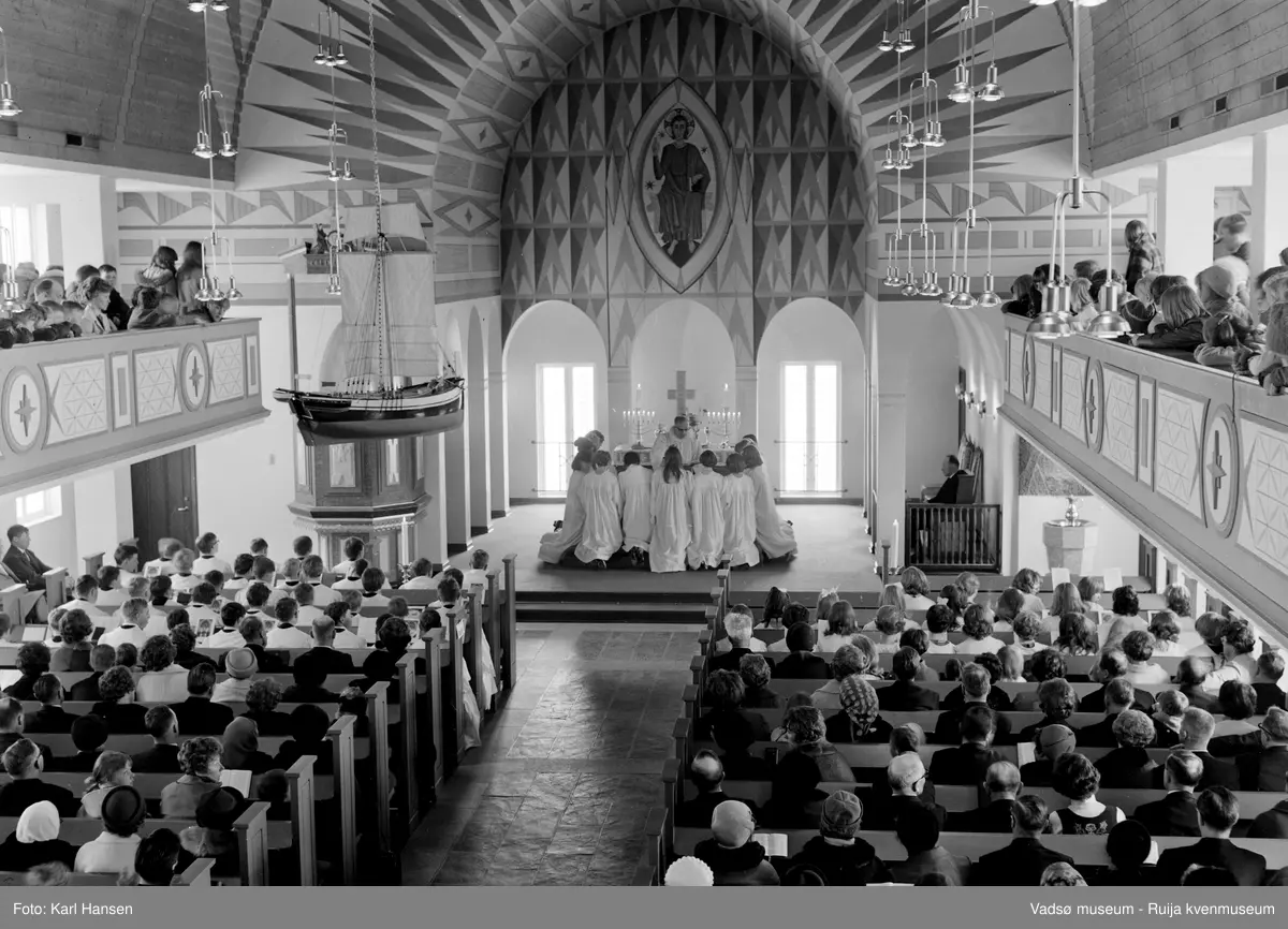 Vadsø kirke, interiør 10.5.1969. Konfirmasjon, konfirmanter kneler ved alteret. Kirka er tegnet av Magnus Poulsson og vigslet i 1958.