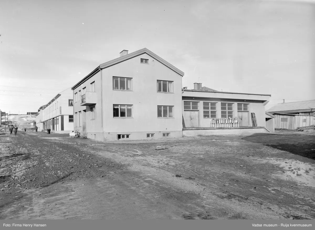 A/L Aust Finnmark Meieri. Bildet er tatt mot nord og viser sydveggen og lasterampen med stabler av melkespann. Det er vårløsning i Tollbugata.  Utenfor meieret står en barnevogn,  sykkel og  spark.
Aust- Finnmark Meieri ble oppført i 1949; arkitekt Monrad Heggen m hovedentreprenør Brdr. Aronsen.
