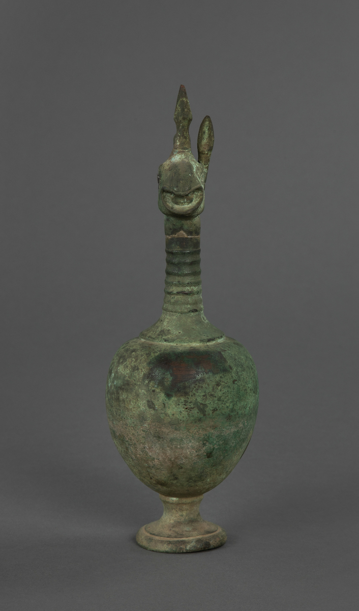 Skulptur av metall. Med en liten stett, hank og fantasidyr hode (fugl/enhjørning). Omtalt som vase men har en veldig grunn åpning i fantasidyrets hode og ut gjennom munnen. Irrgrønn.