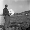 En kepsbeklädd man står på en lerig åker. Han är fotograferad i helfigur. I bakgrunden skymtas ett hus. Han håller en trasig hålyxa i handen.