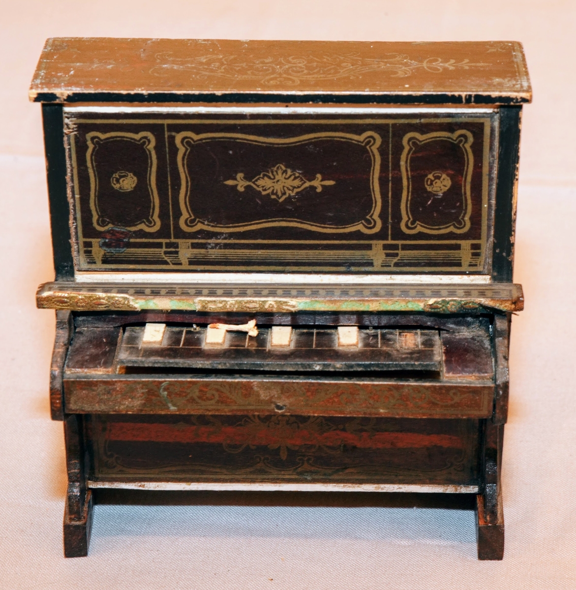 Piano i miniatyr till dockskåp från mitten av 1800-talet. I mörkbrunt målat trä med tryckt dekor i guldig färg på ovansidan och framsidan av pianots ovanstycke och främre nedanstycke. Dekoren föreställer bland annat rocailler, paneler och bårdar i sirlig och asymeterisk stil. Det finns en guldig bård med blomformade maskaroner framtill ovanför tangenterna. Vita fält längst upp och längst ned på främre ovanstycket och nedanstycket (ovanför den tryckta dekoren). Med 15 tangenter (en saknas).