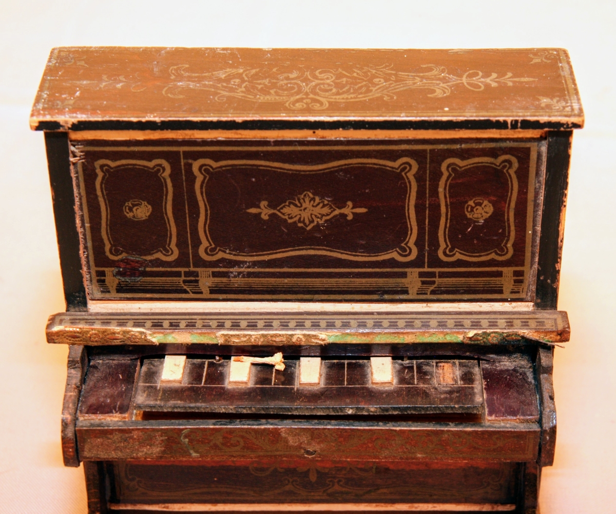 Piano i miniatyr till dockskåp från mitten av 1800-talet. I mörkbrunt målat trä med tryckt dekor i guldig färg på ovansidan och framsidan av pianots ovanstycke och främre nedanstycke. Dekoren föreställer bland annat rocailler, paneler och bårdar i sirlig och asymeterisk stil. Det finns en guldig bård med blomformade maskaroner framtill ovanför tangenterna. Vita fält längst upp och längst ned på främre ovanstycket och nedanstycket (ovanför den tryckta dekoren). Med 15 tangenter (en saknas).