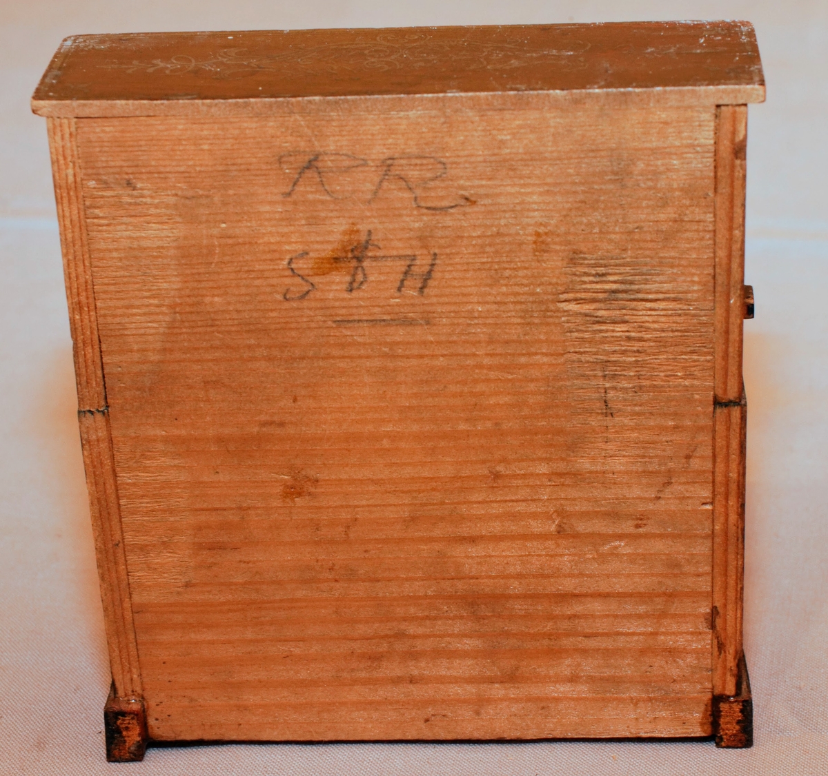 Piano i miniatyr till dockskåp från mitten av 1800-talet. I mörkbrunt målat trä med tryckt dekor i guldig färg på ovansidan och framsidan av pianots ovanstycke och främre nedanstycke. Dekoren föreställer bland annat rocailler, paneler och bårdar i sirlig och asymeterisk stil. Det finns en guldig bård med blomformade maskaroner framtill ovanför tangenterna. Vita fält längst upp och längst ned på främre ovanstycket och nedanstycket (ovanför den tryckta dekoren). Med 15 tangenter (en saknas).