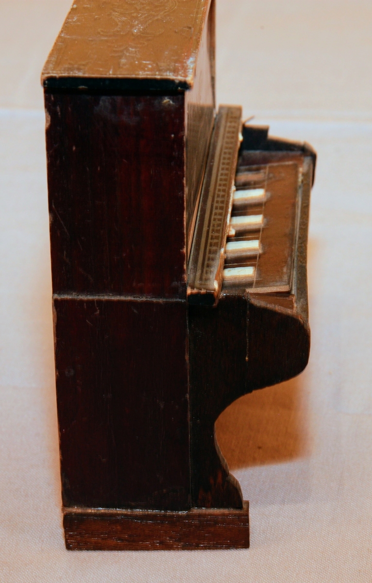 Piano i miniatyr till dockskåp från mitten av 1800-talet. I mörkbrunt målat trä med tryckt dekor i guldig färg på ovansidan och framsidan av pianots ovanstycke och främre nedanstycke. Dekoren föreställer bland annat rocailler, paneler och bårdar i sirlig och asymeterisk stil. Det finns en guldig bård med blomformade maskaroner framtill ovanför tangenterna. Vita fält längst upp och längst ned på främre ovanstycket och nedanstycket (ovanför den tryckta dekoren). Med 15 tangenter (en saknas).