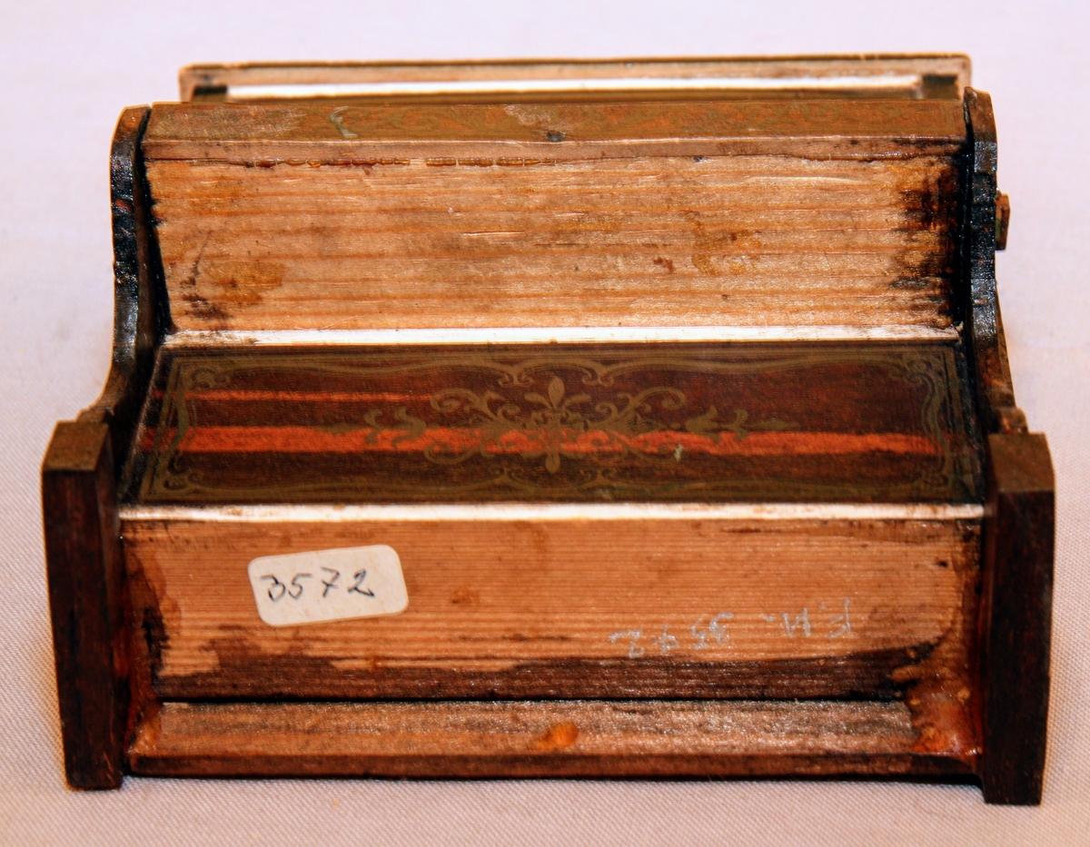 Piano i miniatyr till dockskåp från mitten av 1800-talet. I mörkbrunt målat trä med tryckt dekor i guldig färg på ovansidan och framsidan av pianots ovanstycke och främre nedanstycke. Dekoren föreställer bland annat rocailler, paneler och bårdar i sirlig och asymeterisk stil. Det finns en guldig bård med blomformade maskaroner framtill ovanför tangenterna. Vita fält längst upp och längst ned på främre ovanstycket och nedanstycket (ovanför den tryckta dekoren). Med 15 tangenter (en saknas).