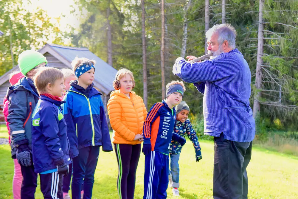 Musiker Stein Villa spiller fløyte, sju 3.-klassinger står rundt og ser på, de ser interesserte og fascinerte ut. Det er et lafta hus og grønn skog i bakgrunnen. 