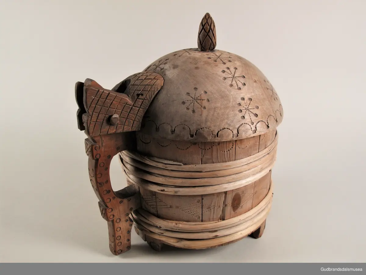 "Ølkrusforma kanne, stor, med kurve lok, som paa undersida har geomegtriske ornament og inskripsjon. Trulig brukt som smørform. Fraa privatsamling..." (Jvf. Katalog...1925).
Stavkanne brukt til servering av smør. Ca. 4,4 liter. 7 gjorder. Hank og hengslet lokk med lokkgrep. Lokket er festet med en tapp gjennom hank og lokkåpneren sine to ører. Lokket dreier seg ved hjelp av tretappen som fungerer som en aksel. Korpus hviler på tre staver, som er lengre enn de øvrige.
Dekor. Lokk med karveskurd og inskripsjoner på innsiden og svidekor på utsiden. Svidekor på korpus og hank. Liten kongle som lokkgrep.