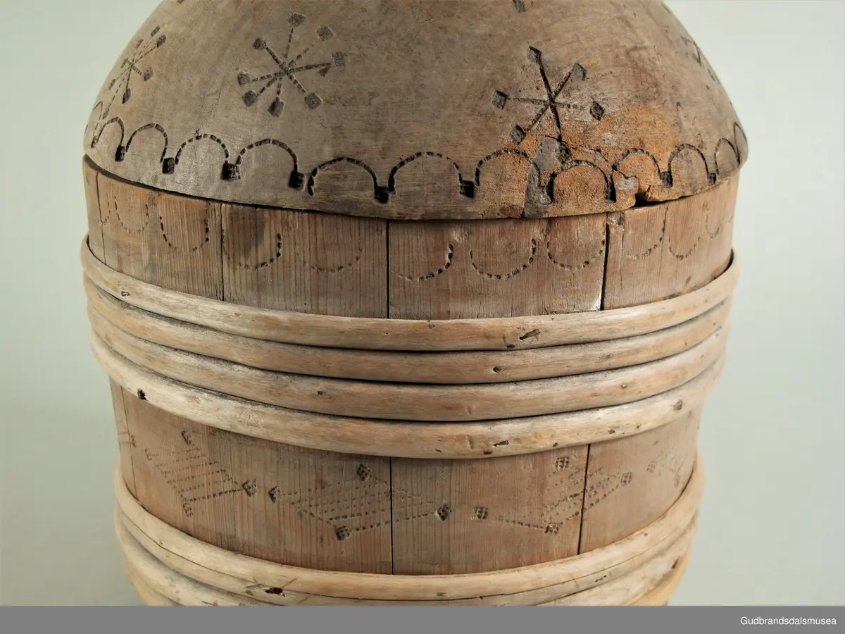 "Ølkrusforma kanne, stor, med kurve lok, som paa undersida har geomegtriske ornament og inskripsjon. Trulig brukt som smørform. Fraa privatsamling..." (Jvf. Katalog...1925).
Stavkanne brukt til servering av smør. Ca. 4,4 liter. 7 gjorder. Hank og hengslet lokk med lokkgrep. Lokket er festet med en tapp gjennom hank og lokkåpneren sine to ører. Lokket dreier seg ved hjelp av tretappen som fungerer som en aksel. Korpus hviler på tre staver, som er lengre enn de øvrige.
Dekor. Lokk med karveskurd og inskripsjoner på innsiden og svidekor på utsiden. Svidekor på korpus og hank. Liten kongle som lokkgrep.