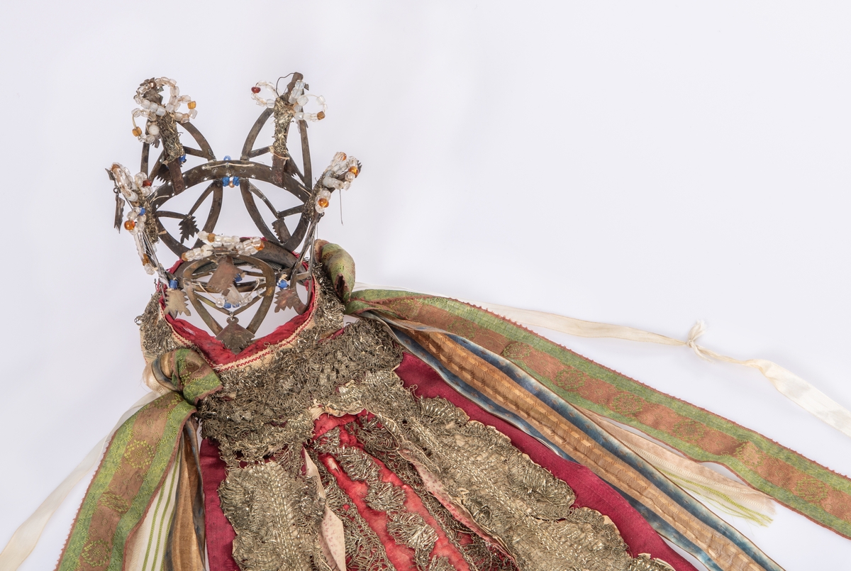 Brudekrone i metall med fem spir. Krona er pyntet med metallheng og glassperler. Det er påsydd valk nederst, med brodert bånd og metallknipling rundt valken.

Bak på det broderte båndet rundt valken, er det festet bånd. På midten er det et bredt bånd i rødt med metallkniplinger, med brede bånd med lys bunn og med en rød kant vendt ut fra den røde midtbåndet. Metallkniplinger er festet til de tre brede båndene. Deretter fire smalere bånd på hver side av de brede båndene. Disse er festet samlet med en liten sløyfe på det bredeste der det er festet på valken. 

En plastkam er påsydd valken sammen med to hvite, nyere bånd. Antagelig gjort i nyere tid for å feste krona og knyte under haka.