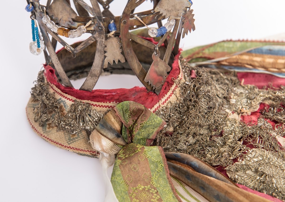Brudekrone i metall med fem spir. Krona er pyntet med metallheng og glassperler. Det er påsydd valk nederst, med brodert bånd og metallknipling rundt valken.

Bak på det broderte båndet rundt valken, er det festet bånd. På midten er det et bredt bånd i rødt med metallkniplinger, med brede bånd med lys bunn og med en rød kant vendt ut fra den røde midtbåndet. Metallkniplinger er festet til de tre brede båndene. Deretter fire smalere bånd på hver side av de brede båndene. Disse er festet samlet med en liten sløyfe på det bredeste der det er festet på valken. 

En plastkam er påsydd valken sammen med to hvite, nyere bånd. Antagelig gjort i nyere tid for å feste krona og knyte under haka.